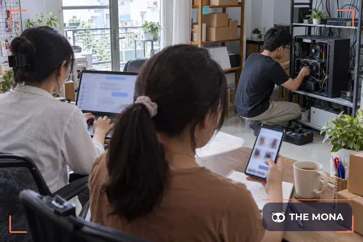Chủ shop xem giỏ hàng trên điện thoại từ phía sau vai, bên cạnh là laptop mở trang quản lý